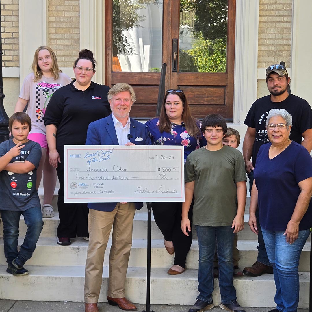 Mayor Gibson holds a big check for a presentation of the June 2024 First Place Winner on July 29th, 2024. Jessica Odom is shown with her family and Valencia Hall, Alderwoman Ward 5 of Natchez. They are standing on the steps in front of City Hall.